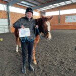 Young adult, Ben stands next to his horse, holding a sign that says "Autism Community Impact Grant"